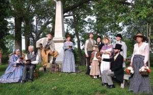A group of people in a cemetery in front of an obelisk monument who are all dressed in 1850s attire.