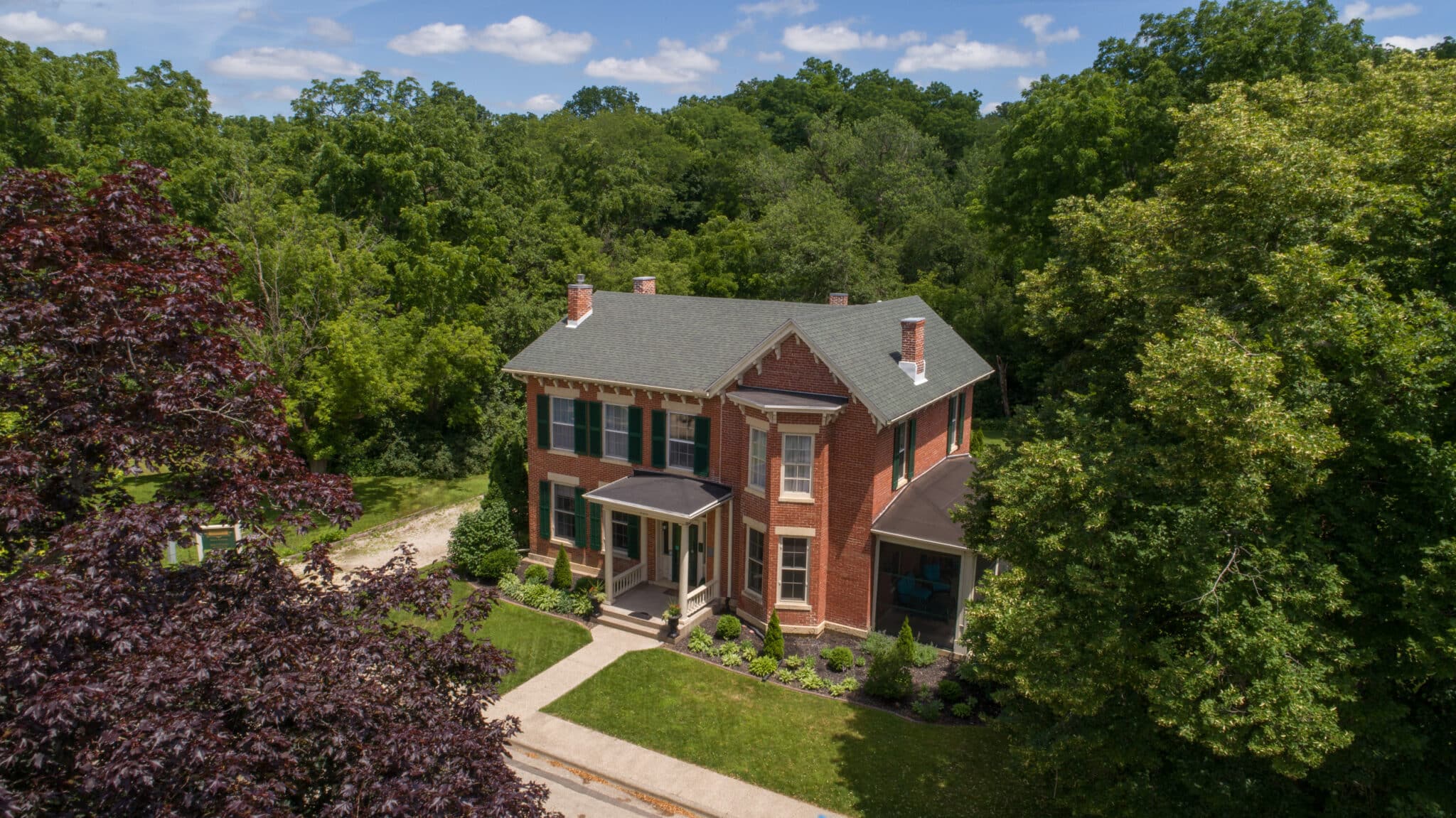 Aerial view of a brick 2-story house with lush green and purple trees and blue skies speckled with a few clouds.