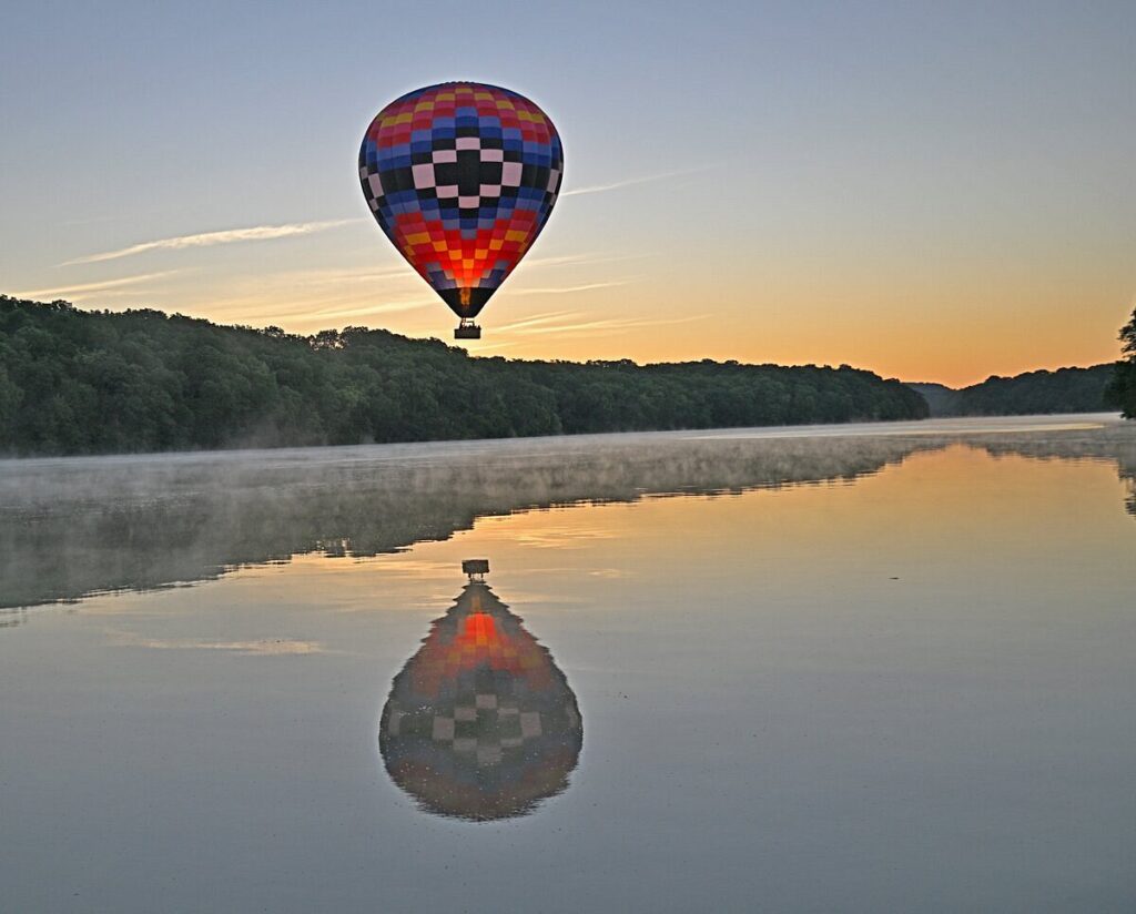 Colorful hot air balloon flies low over a lake surrounded by bluffs with trees