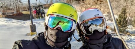 Two skiers with helmets and goggles riding up on a chair lift taking a selfie with other skiers and a river in the background