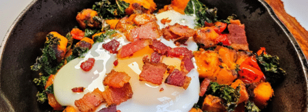 bacon pieces on top of a shirred egg on top of sweet potatoes, kale, and sweet red peppers in a cast iron skillet