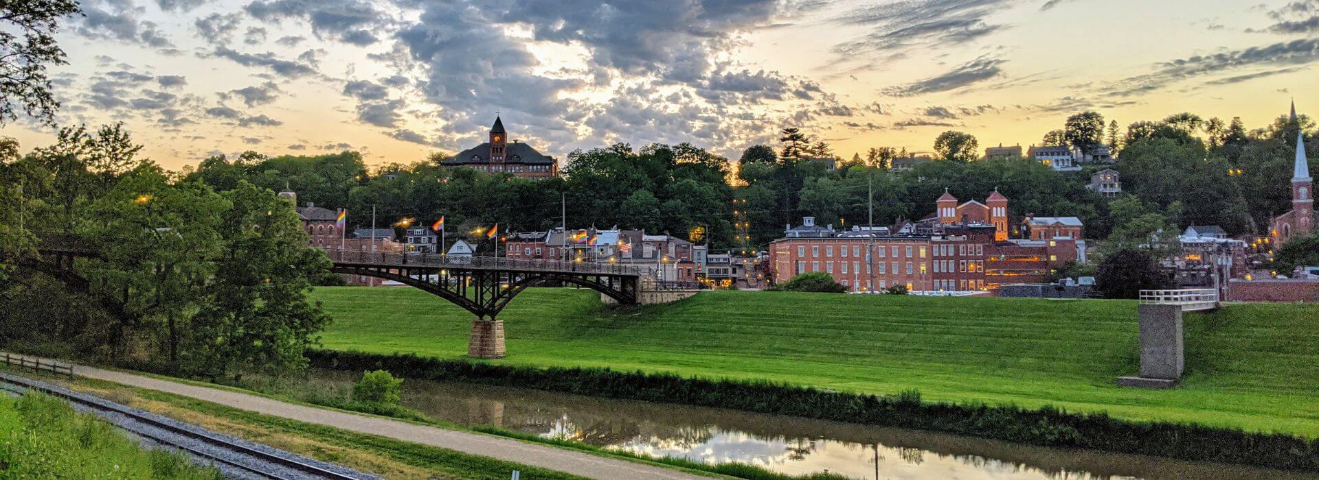 Outdoor picture of a river, green grass, a bridge, brick buildings, and green trees.