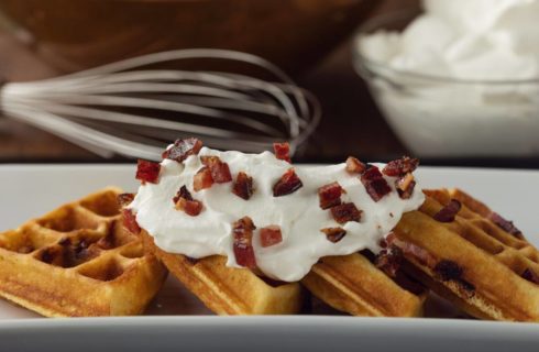 Crispy waffles topped with delicate whipped cream and pieces of bacon with a whisk and bowl of whipped cream in the background.