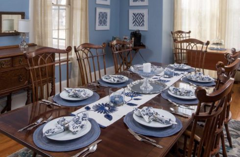 Dining room with blue walls, a wooden dining table and chairs, with a white and blue table runner, round blue placemats with white dishes, white and blue cloth napkins, and silverware. An antique wood side table with mirror along the wall, and a small folding table and chairs along the windows.