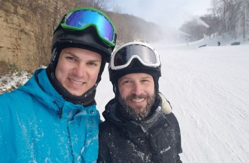 Two men outside on a snowy day wearing blue and black parkas and ski helmets with goggles.