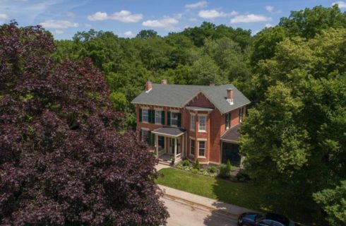 Aerial view of a brick 2-story house with lush green and purple trees and blue skies speckled with a few clouds.
