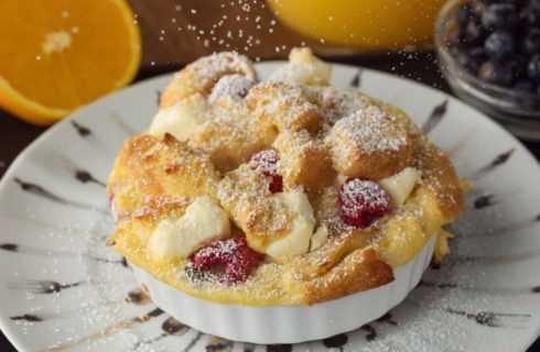 Golden bread pieces baked in a white ramekin with fresh raspberries sprinkled with powdered sugar, on a white plate with black decor, and oranges and fresh blueberries in the background.