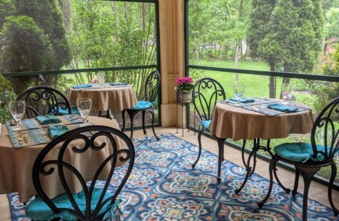 Screened in porch with dining tables covered with light brown tablecloths and blue napkins, glassware, and iron chairs with teal cushions, with views of lush greenery outside.