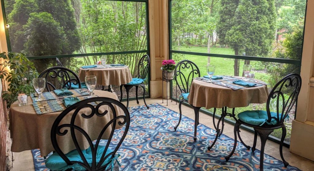 Screened in porch with dining tables covered with light brown tablecloths and blue napkins, glassware, and iron chairs with teal cushions, with views of lush greenery outside.