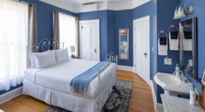bedroom with wood floors, blue walls, white doors, ceiling and trim, a blue and tan tapestry area rug, a white wrought iron bed with white and blue bedding, a round wooden nightstand with white lamp next to the bed, a white pedestal sink with a rectangular mirror above it, and white and black towels next to the sink, along with tall windows with white and tan curtains.