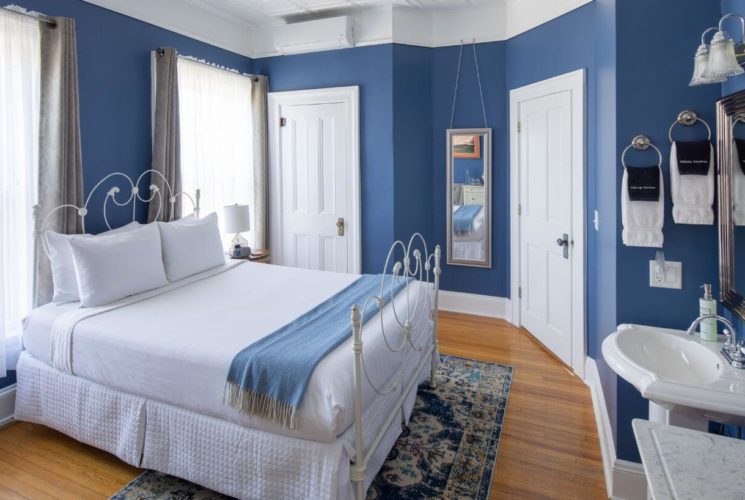 Room with wood floors, bed with white wrought iron head and foot scroll, white linens on bed with blue throw, blue walls with white doors, trim and ceiling.