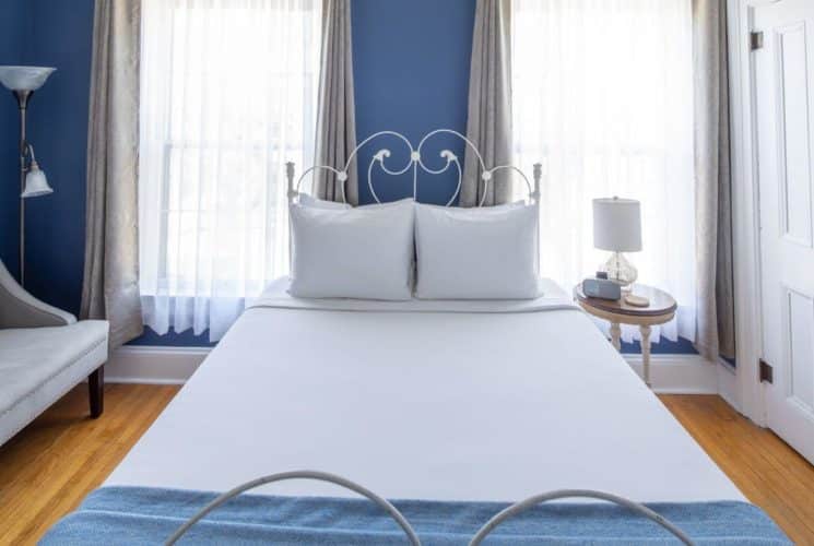 bedroom with wood floors, blue walls, white door and trim, a grey couch with a tall lamp next to the couch, a white wrought iron bed with white and blue bedding, a round wooden nightstand with white lamp and alarm clock