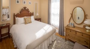 room with yellow walls, bed with antique carved wood headboard, white linens, and a yellow, grey and white mandala throw pillow, wood nightstands with lamps on either side of bed, wood dresser with mirror, an upholstered armchair, and wood floors with a tapestry rug