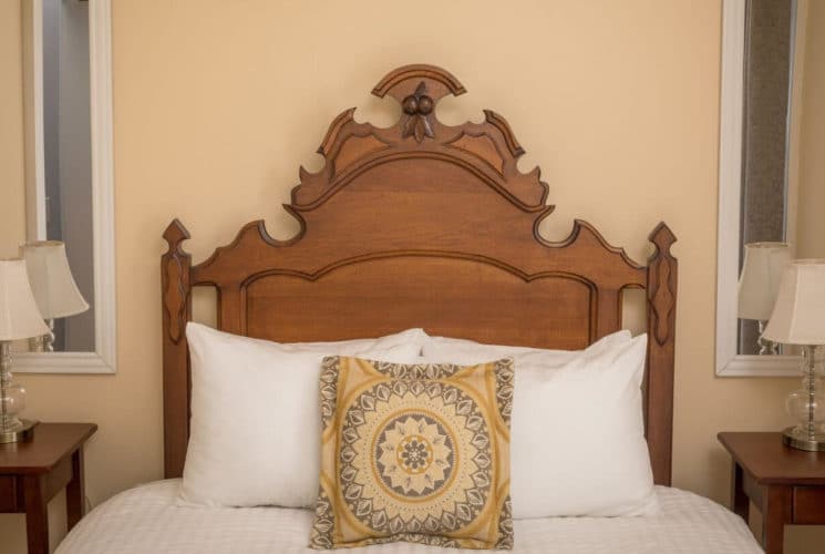 close up of wooden antique carved headboard, white linens on the bed, along with a yellow and gray mandala pillow, and 2 nightstands on either side of the bed with lamps, and mirrors behind the lamps.