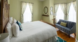 Bedroom with light sage green walls, bed with ornate wood headboard, white bedding on the bed with teal and white throw pillow, teal and white run, wood floors, a blue sofa, a side table with lamp, and windows with cream and green curtains.