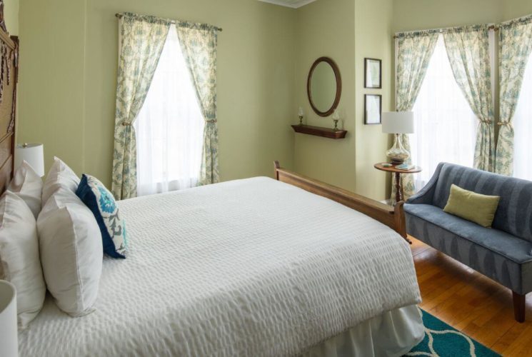 Bedroom with light sage green walls, bed with ornate wood headboard, white bedding on the bed with teal and white throw pillow, teal and white run, wood floors, a blue sofa, a side table with lamp, and windows with cream and green curtains.