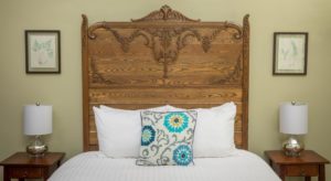 Close up of detailed carved wood headboard, bed with white bedding and white, gray and blue throw pillow, wooden nightstands on either side of the bed with lamps.