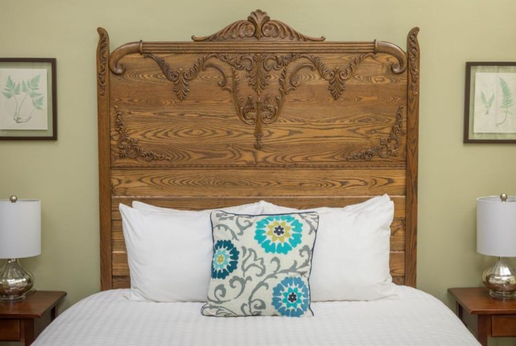 Close up of detailed carved wood headboard, bed with white bedding and white, gray and blue throw pillow, wooden nightstands on either side of the bed with lamps.