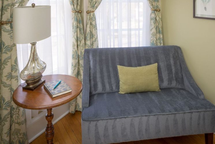 corner of room with blue oversized chair, wood side table with lamp, wood floors, yellow walls, and windows with sheer curtains and beige, green and blue curtains.