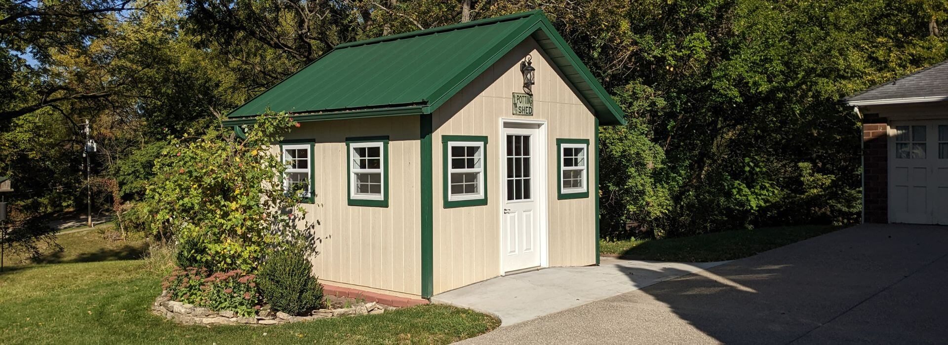 tan shed with green roof and trim, and a white door, with green trees in the background and a sidewalk and green grass.