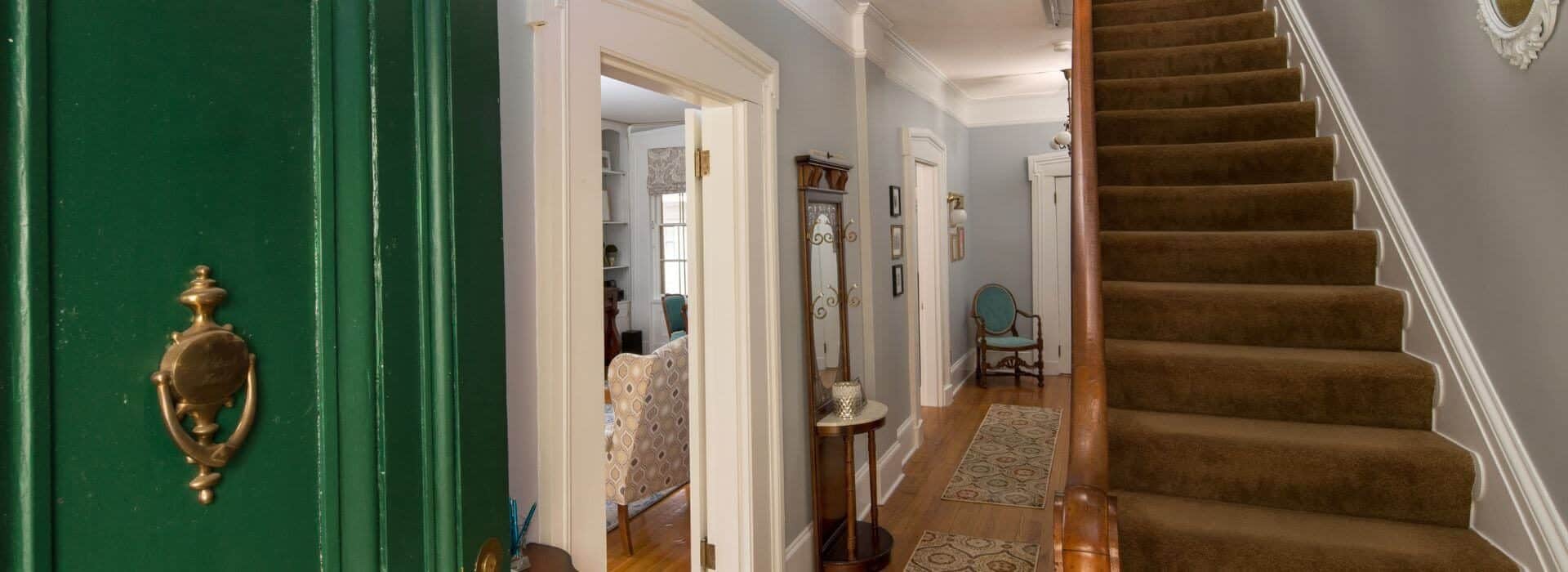 Foyer with open green door with brass knocker, stairway with carpeting, gray walls with white trim, wood floor with tapestry rugs, and anitique furniture