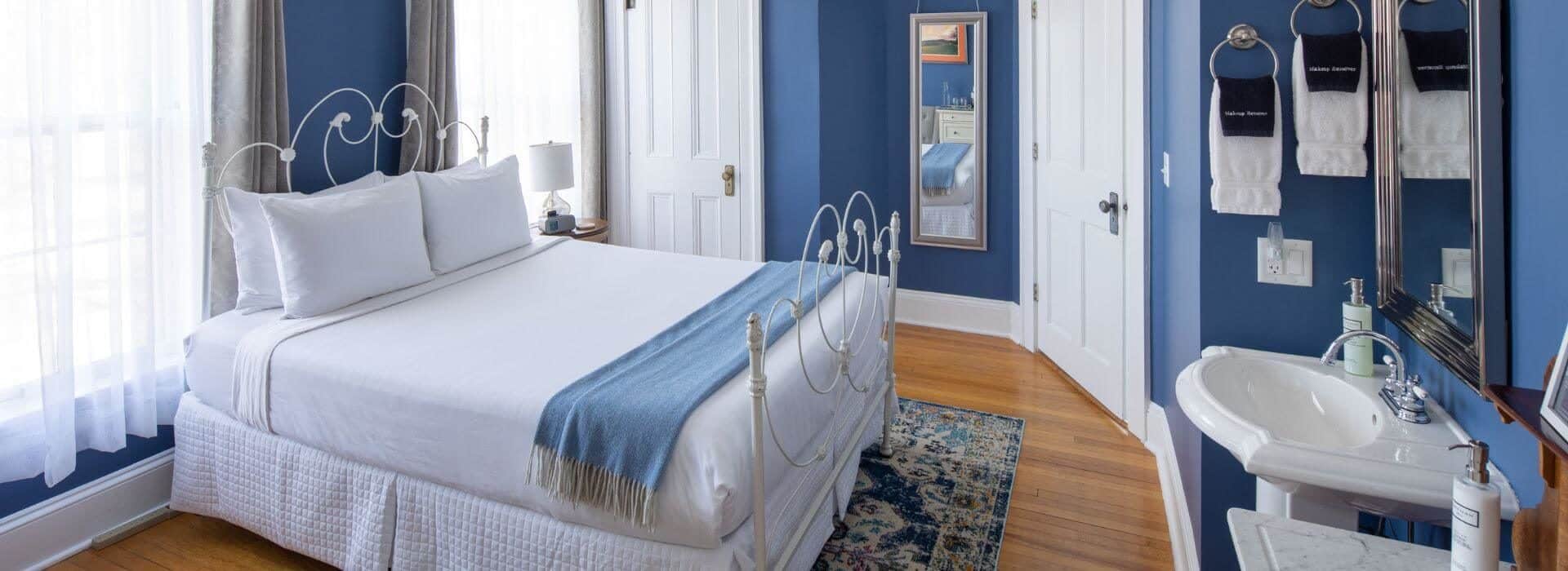 Bedroom with wood floors, blue walls with white trim and doors, a wrought iron bed with white and blue linens, a tapestry rug, and pedestal sink with mirror.