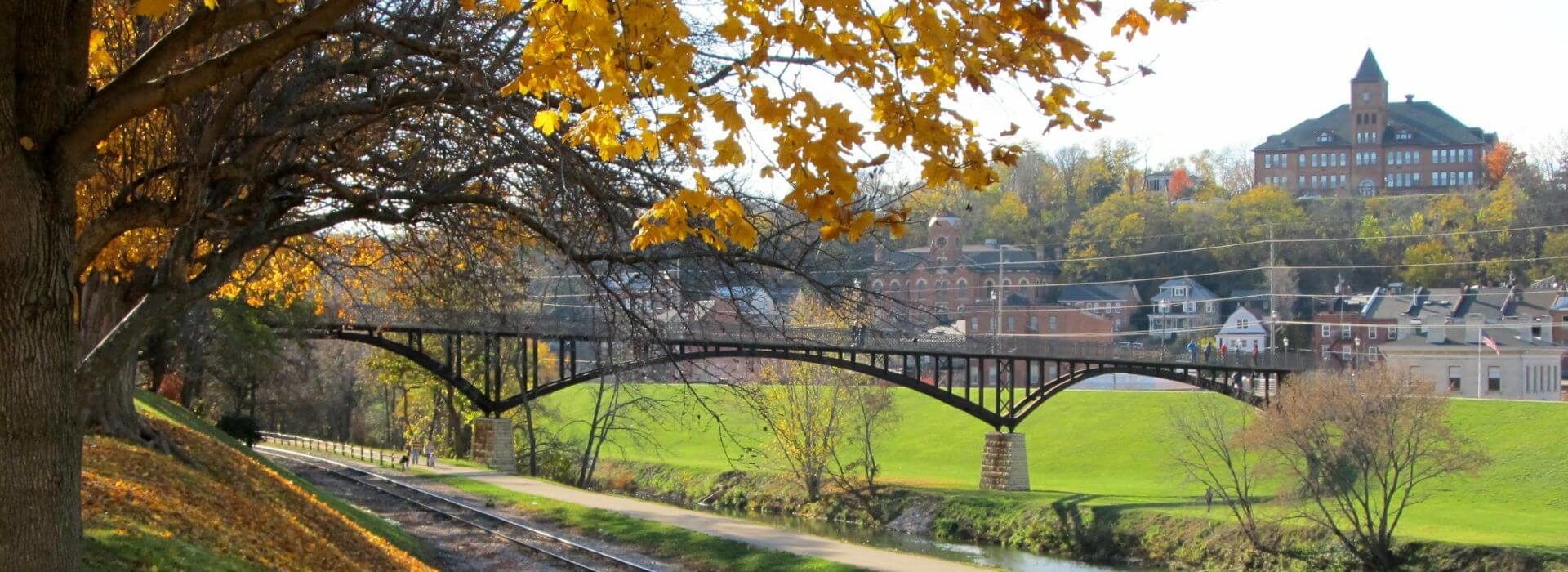 Landscape of green grass a river, a bridge, and a tree with fall foliage, and buildings in the background