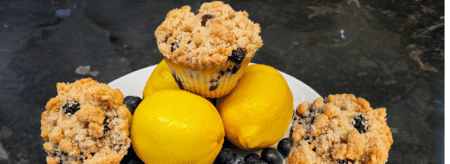 three lemon blueberry muffins on a white plate surrounded by whole lemons and blueberries