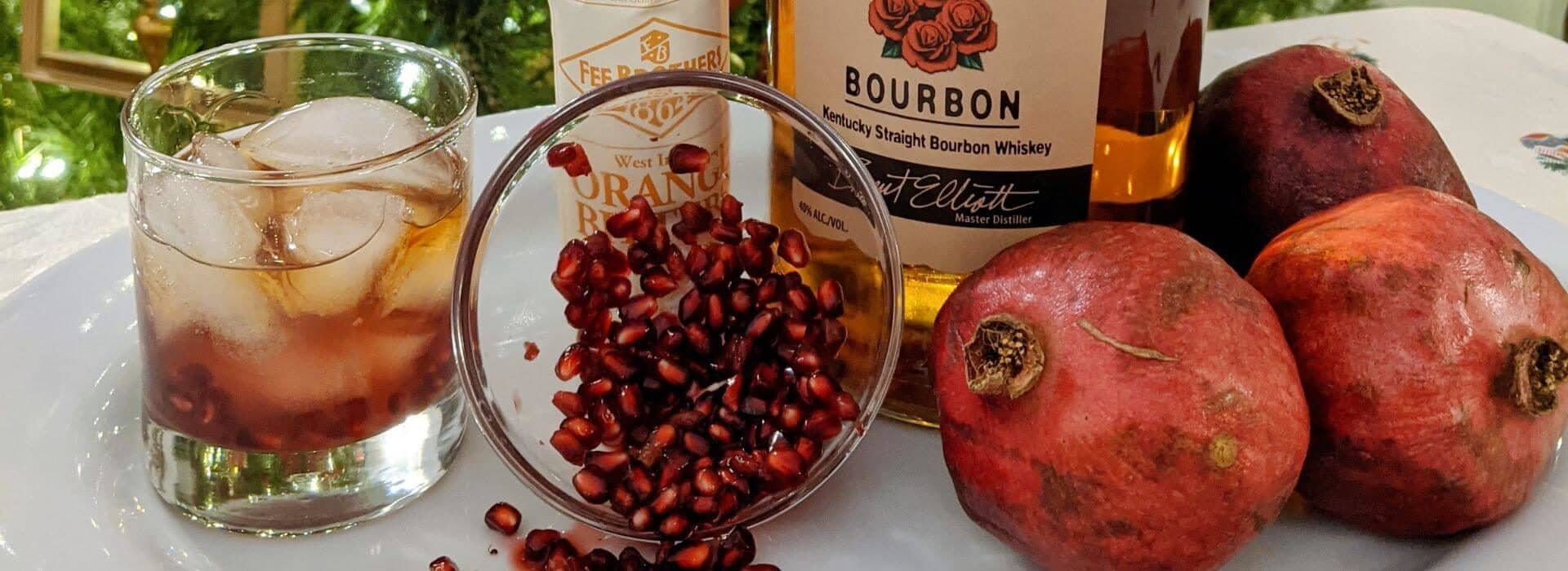a spread of whole red pomegranates, a glass full of pomegranate seeds, bottles of bourbon and orange liqueur, and a clear glass full of ice and an amber colored beverage.