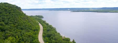 An aerial view of a winding road with a river on the right and tree covered bluffs on the left