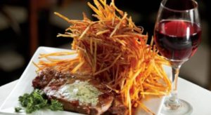 White plate with steak covered with herb butter, adorned with piles of thinly shredded pieces of potato fries, garnished with parsley and accompanied by a glass of red wine.