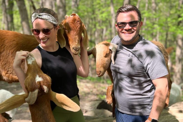 man and woman wearing sunglasses, surrounded by goats with trees in the background